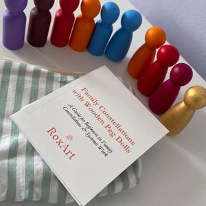 May include: A collection of colorful wooden peg dolls in various colors, including purple, brown, red, orange, blue, and gold. A book titled "Family Constellations with Wooden Peg Dolls" is also present, along with a striped pouch.