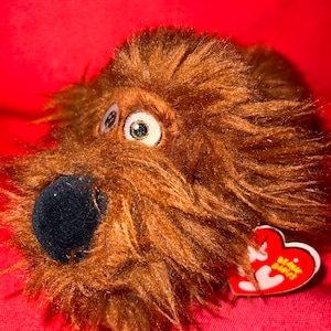 May include: A brown plush dog toy with a black nose and large, expressive eyes. The toy has a shaggy fur texture and a red heart-shaped tag that reads "Beanie Babies". The toy is set against a red background.