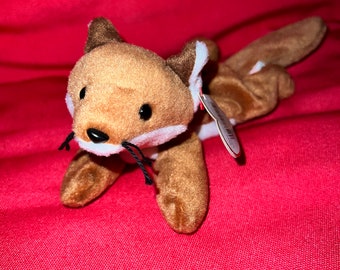 Puede incluir: Un peluche de zorro con pelaje marrón, detalles blancos y bigotes negros. El zorro tiene una etiqueta adjunta. El juguete está sobre un fondo rojo.