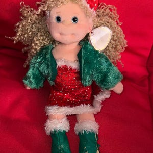 May include: A plush doll with blonde curly hair, blue eyes, and a red bow. The doll wears a red dress with white accents, a green jacket, and green boots. The doll is a Christmas-themed collectible.