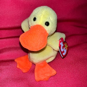 May include: A yellow plush duck toy with a bright orange beak and orange webbed feet. The stuffed animal features black plastic eyes and a red heart-shaped tag attached to its side with the white letters ty and the words Beanie Baby printed.