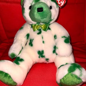 May include: A white plush bear with green shamrock patterns and accents. The bear has a green bow tie, ears, and nose. A red heart-shaped tag is attached to the ear. The bear is sitting on a red surface.