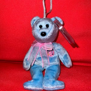 May include: A blue plush beanie baby bear with a pink ribbon and a heart-shaped tag. The bear is sitting on a red surface.