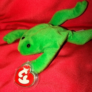 Könnte beinhalten: Ein leuchtend grünes Frosch-Stofftier mit schwarzen Augen, das an einem der Vorderbeine ein herzförmiges, rot-goldenes Ty-Etikett mit einer durchsichtigen Kunststoffhülle trägt. Das Plüschtier hat eine weiche, flauschige Textur und einen flachen Körper.