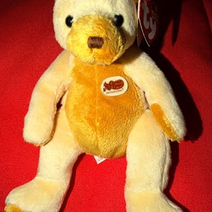 May include: A plush bear toy with cream and golden-yellow fur, featuring black plastic eyes and a brown nose. An oval patch on the chest displays the text Cracker Barrel Old Country Store. A red heart-shaped Ty Beanie Babies tag is attached.