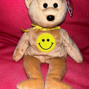 May include: A tan plush teddy bear with a yellow smiley face on its chest and a yellow ribbon around its neck. The bear has black eyes and a black nose. A red tag is attached to its ear.