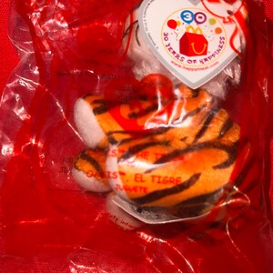 Puede incluir: Un pequeño tigre de peluche en una bolsa de plástico transparente. El tigre tiene rayas naranjas y negras con detalles blancos. Una etiqueta en forma de corazón dice "30 Years of Happiness" y "Happy Meal". El texto "Oasis, El Tigre, Juguete" es visible.