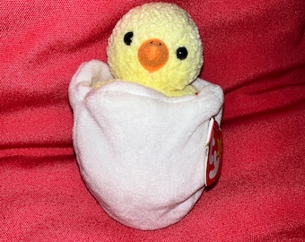 Peut inclure: Une peluche représentant un petit poussin jaune à la texture bouclée sortant d'une coquille d'œuf blanche et lisse. Le poussin possède deux yeux en perles noires et un bec orange. Une étiquette Ty rouge en forme de cœur est fixée sur le côté.