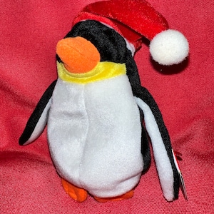 May include: A plush penguin toy with a red Santa hat and a white pom-pom. The penguin has a black head and wings, a white body, and an orange beak and feet. A yellow scarf is around its neck. The toy is on a red background.