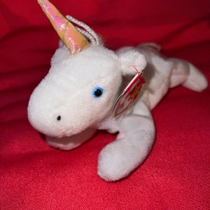 Puede incluir: Unicornio de peluche blanco con un cuerno rosa e iridiscente. El unicornio tiene ojos azules y una etiqueta roja de Beanie Baby unida a su pierna.