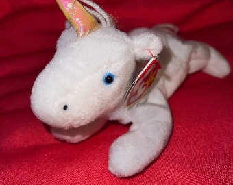 Könnte beinhalten: Weißes Plüsch-Einhorn mit einem rosa und schillernden Horn. Das Einhorn hat blaue Augen und ein rotes Beanie Baby-Etikett an seinem Bein.