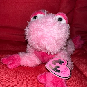 Könnte beinhalten: Ein rosa Plüschfrosch-Spielzeug mit einem herzförmigen Anhänger. Der Frosch hat große schwarze Augen, rosa Akzente und eine weiche, flauschige Textur. Auf dem Anhänger steht "Pinky" und "Beanie Babies".