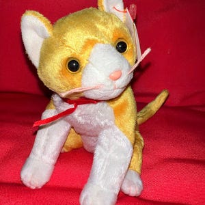May include: A yellow and white plush cat with a red ribbon around its neck. The cat is sitting on a red surface.