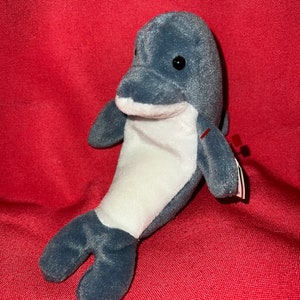 Puede incluir: Un juguete de peluche de delfín gris y blanco. El delfín tiene la barriga blanca y la espalda gris con un ojo negro. El juguete tiene una etiqueta roja adjunta.