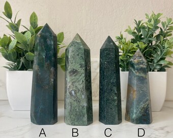 May include: Four green moss agate crystal towers with pointed tops. The crystals have varying shades of green and brown, with visible patterns. The crystals are labeled A, B, C, and D. The crystals are displayed in front of a white background with green plants.
