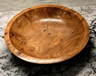 May include: A handcrafted wooden bowl with a natural wood grain pattern. The bowl is a warm brown color and has a smooth, polished finish. The bowl is round with a wide, shallow design.