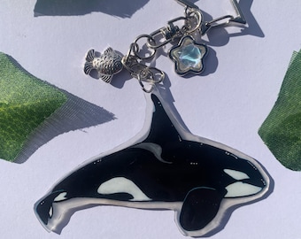 May include: A clear acrylic keychain featuring a black and white orca whale illustration. The keychain includes a silver-toned metal star-shaped clasp, a small silver fish charm, and a small star charm with a pearlescent, iridescent finish.