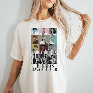 May include: A white t-shirt with a graphic print of the cast of Grey's Anatomy. The graphic is a collage of photos of the actors in different colored squares. The text "The Greys Rewatch Tour" is printed below the graphic.