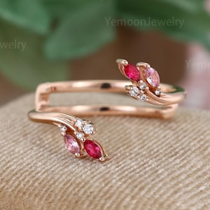 May include: Rose gold ring with a bypass design. The ring features marquise-cut pink and red gemstones, accented with small white gemstones. The ring is set on a beige surface.