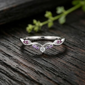 May include: A silver-toned V-shaped ring featuring a central clear round-cut stone flanked by four small purple stones set into the band on each side, creating a symmetrical design that tapers toward the ends of the band.