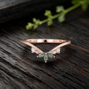 May include: A V-shaped rose gold ring featuring a central cluster of three marquise-cut green stones arranged in a leaf-like pattern, flanked by two small round clear stones set into the band, which displays a delicate engraved leaf texture.