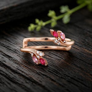 May include: A rose gold open ring featuring two bypass ends, each adorned with marquise-cut pink and red gemstones and small round clear stones. The band has a polished finish and a split design that curves around the finger for a layered look.