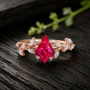 May include: A rose gold ring featuring a kite-shaped vibrant red gemstone as the center stone. The band is designed with a vine-like motif, adorned with multiple small marquise-cut white stones arranged to resemble leaves along the sides.