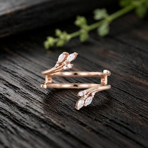 May include: A rose gold open ring featuring two leaf-shaped ends, each adorned with small, clear, marquise-cut and round-cut stones. The band is smooth and polished, with the two ends curving toward each other without meeting in the center.