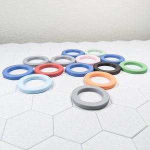 May include: A collection of 12 colorful plastic rings, arranged in a circular pattern on a white hexagonal tile surface. The rings are in various colors, including blue, red, green, orange, pink, and gray.