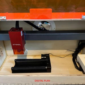 以下が含まれることがあります： 赤と黒のレーザーカッターで、黒い金属ビームと白いコントロールパネルが付いています。レーザーカッターは、赤い天蓋が付いた木製の箱の中にあります。箱には、底に「DIGITAL PLAN」という文字が書かれています。