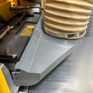 May include: A close-up of a woodworking machine with a gray dust collection port and a flexible beige hose. A wrench and a warning label are visible on the machine's black surface. The machine has a yellow frame.
