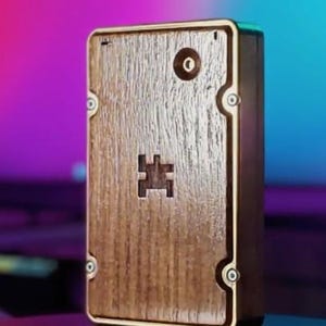 May include: A rectangular wooden box with a light brown finish and a gold-colored frame. The box features a small, square-shaped cutout and a circular detail on the front. The wood grain is visible, adding texture to the design. The box is likely for storage.