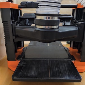May include: A portable thickness planer featuring a black and orange frame, a flat black metal feed table, and a flexible white dust collection hose attached to the top center port with a metal clamp. The machine has a top adjustment knob.