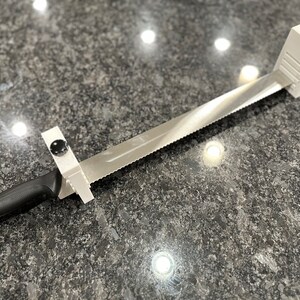 以下が含まれることがあります： 黒いノブが付いた白いプラスチック製のパンナイフガイドと、黒いハンドルが付いた鋸歯状のステンレス製のパンナイフです。