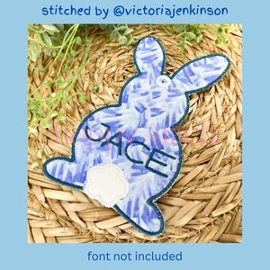 May include: A blue and white patterned bunny-shaped applique with the name "JACE" embroidered on it. The applique has a white fluffy tail and a dark blue border. Text at the top reads "stitched by @victoriajenkins", and at the bottom, "font not included".