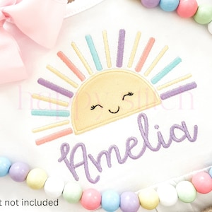 May include: A white embroidered design of a rainbow sun with a smiling face and the name Amelia in purple cursive lettering. The design is on a white fabric background with a pink bow and pastel colored beads around the edges.