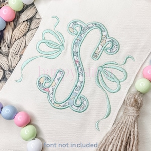 May include: A light green embroidered letter S with a floral pattern inside the letter. The letter is on a white fabric background with a brown tassel.