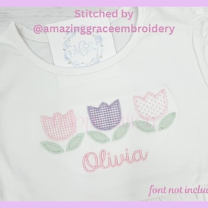 May include: White t-shirt with a floral design featuring three embroidered tulips in pink, purple, and white. The name "Olivia" is embroidered below the flowers. The text "Stitched by @amazinggraceembroidery" is at the top.