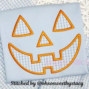 May include: A light blue square with an embroidered orange jack-o'-lantern face. The face has triangular eyes, a triangular nose, and a smiling mouth. The text "Stitched by @ohsosweetbystacy" is at the bottom.