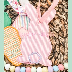 May include: An Easter-themed decorative tag featuring a pink and white striped bunny shape with a floral ribbon. Other tags include a green shape and an orange gingham tag with the word "FUN". The background is woven wicker.