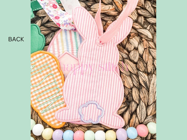 May include: An Easter-themed decorative tag featuring a pink and white striped bunny shape with a floral ribbon. Other tags include a green shape and an orange gingham tag with the word "FUN". The background is woven wicker.