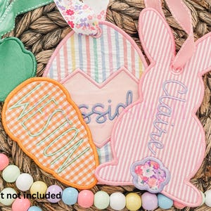 May include: Embroidered Easter-themed gift tags in the shape of a carrot, egg, and bunny. The carrot tag is orange and white checkered, the egg tag is striped, and the bunny tag is pink and white striped. Each tag has a name embroidered on it.