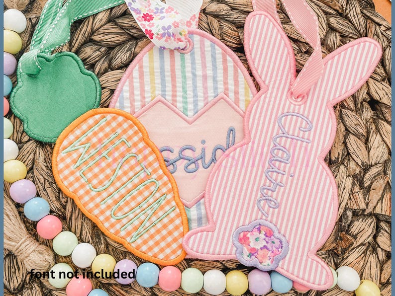 May include: Embroidered Easter-themed gift tags in the shape of a carrot, egg, and bunny. The carrot tag is orange and white checkered, the egg tag is striped, and the bunny tag is pink and white striped. Each tag has a name embroidered on it.