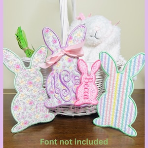 May include: Embroidered bunny-shaped decorations in floral, striped, and monogrammed designs. A pink bunny with the name "Becca" is also present. A white basket holds a stuffed animal and green decorative grass.