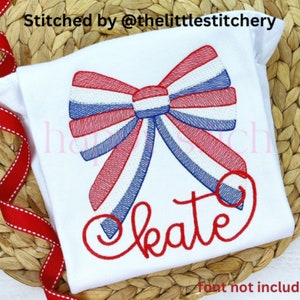 May include: A white t-shirt with a red, white, and blue bow embroidered on it. The word "kate" is embroidered below the bow in red thread. "Font not included" is printed below the design.