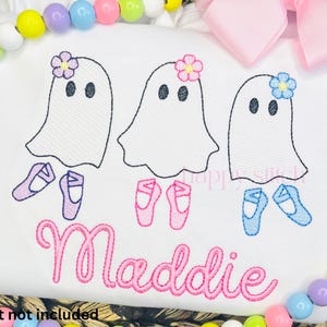 May include: White t-shirt featuring three ghost designs, each adorned with a flower and ballet shoes. The name "Maddie" is embroidered in pink script below the ghosts. A pink bow and colorful beads decorate the border.