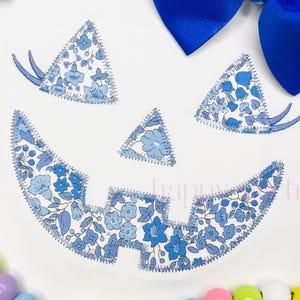 Puede incluir: Un fondo blanco presenta un diseño floral azul con forma de cara de calabaza. El diseño está hecho de tela con un estampado floral y bordes cosidos. Un gran lazo azul en la parte superior. La palabra "happy" es visible.