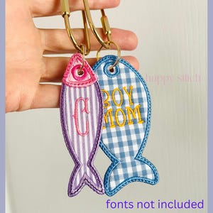 May include: Two fish-shaped embroidered keychains attached to a gold-tone metal carabiner. The left keychain features purple and white stripes with a pink embroidered letter C. The right keychain has a blue and white gingham pattern with yellow text reading BOY MOM.
