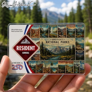 May include: A clear plastic Resident Annual America 250 National Parks pass. The pass features illustrations of various national parks, including Yosemite, Yellowstone, and the Grand Canyon. The pass is held in front of a blurred natural background.