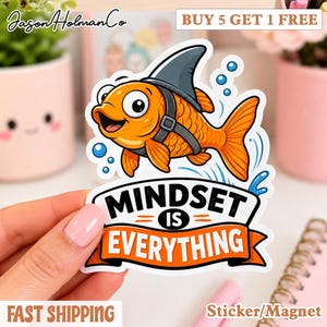 May include: A die-cut sticker featuring an orange goldfish wearing a gray shark fin harness, surrounded by blue bubbles. Below the fish, a banner contains the black and orange text "MINDSET IS EVERYTHING" in a bold, sans-serif font.